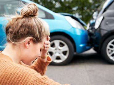 Female Motorist With Head In Hands Sitting Next To Vehicles Involved In Car Accident